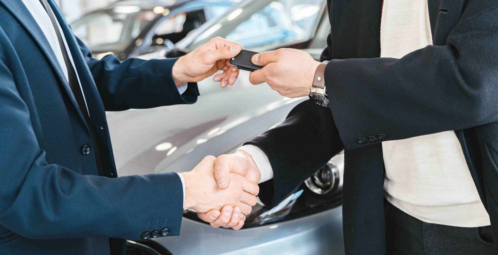 Two People Shacking Hands And One Passing Car Keys To The Other In Front Of A Vehicle